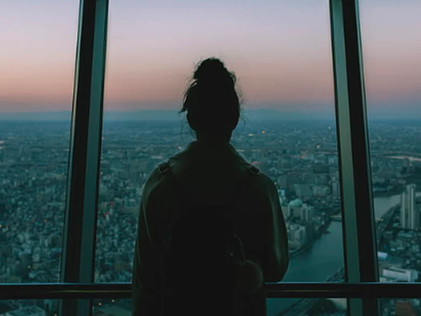 Mulher de costas olhando uma grande cidade ao entardecer por uma janela panorâmica