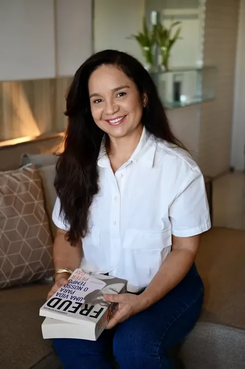 Mônica Veloso, psicanalista, sorrindo no consultório com um livro sobre Freud nas mãos