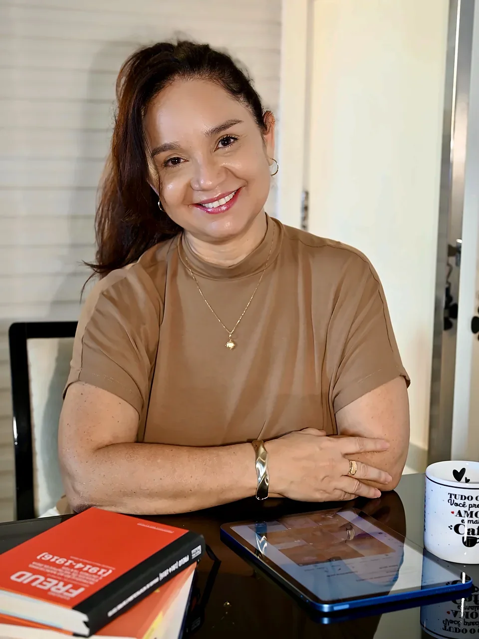 Mônica Veloso, psicanalista, sorrindo à mesa do consultório com livros de Freud, tablet e caneca de café